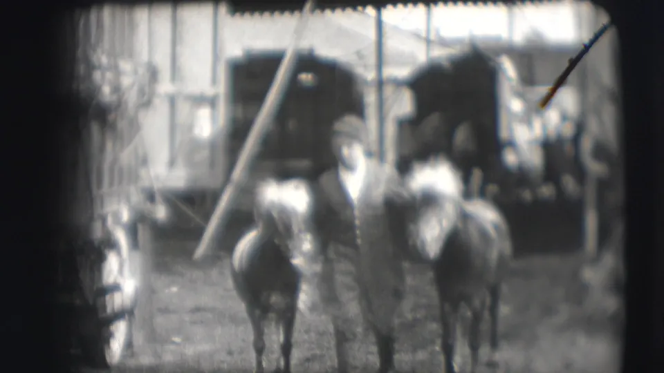 Soft black-and-white frame shows a man and two horses moving near circus wagons, poles, and canvas structures in the 1948 Sarasota setting.
