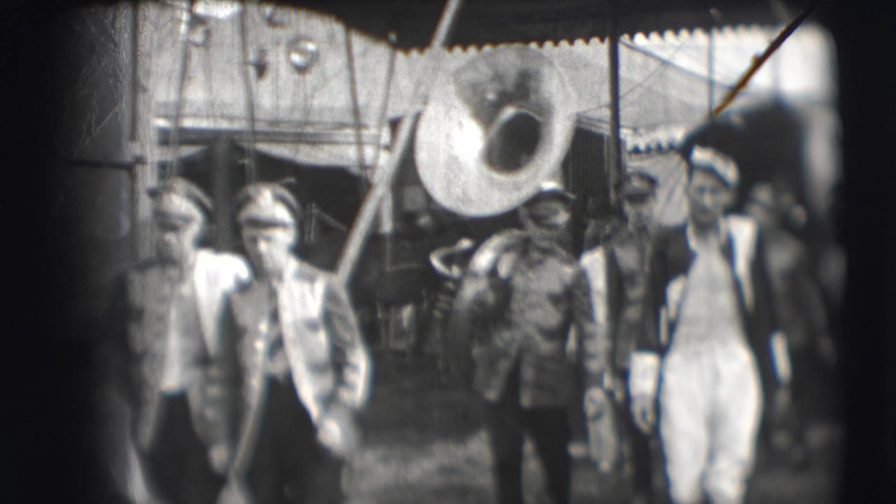 Several men walk under a tented area, including uniformed figures and a large brass instrument.