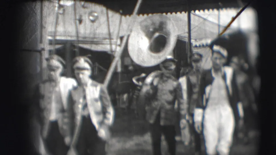 Men in uniform and civilian clothing walk through the Ringling Brothers Sarasota circus area, with a large brass instrument overhead.