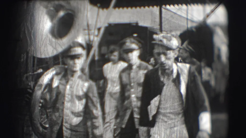 In the Sarasota circus setting, uniformed men and an older bearded man walk toward the camera beneath tent rigging and a brass instrument.