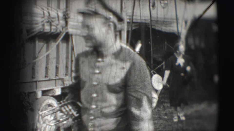 A uniformed circus musician walks close to the camera with a brass instrument near wagon boards and hanging rigging in Sarasota footage.