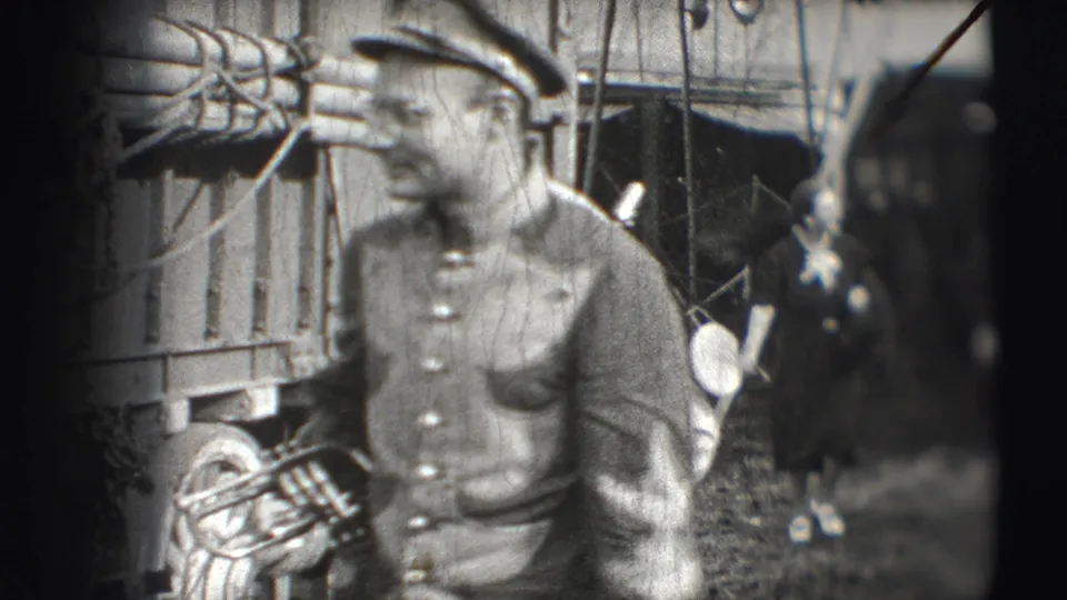 The 8mm frame shows a close, soft-focus view of a uniformed circus musician holding a brass instrument beside a wagon.