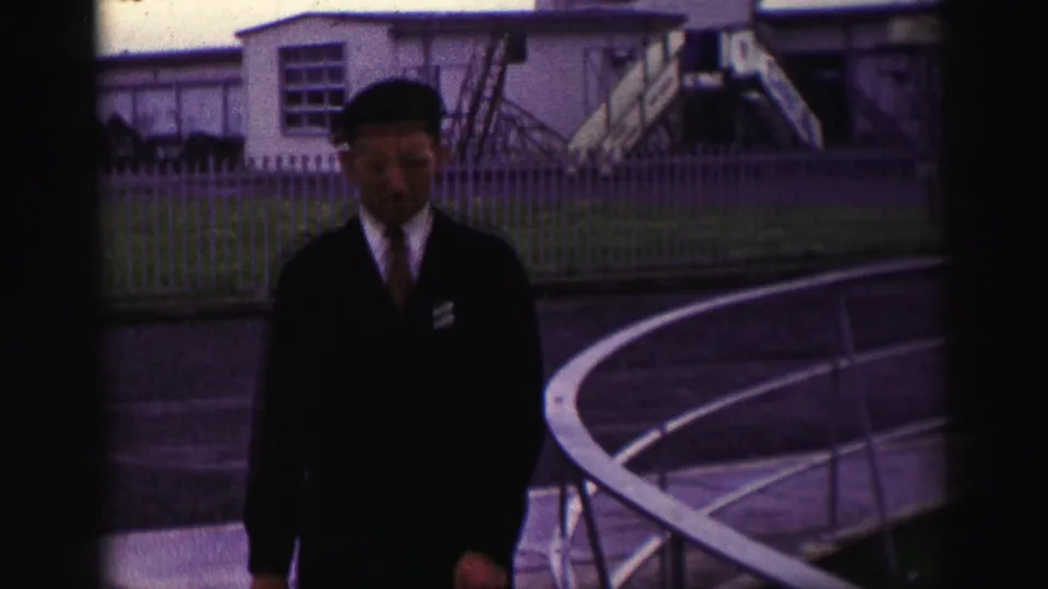 1952 United Kingdom home-movie frame showing a uniformed man in a beret walking near railings with low base buildings behind him.