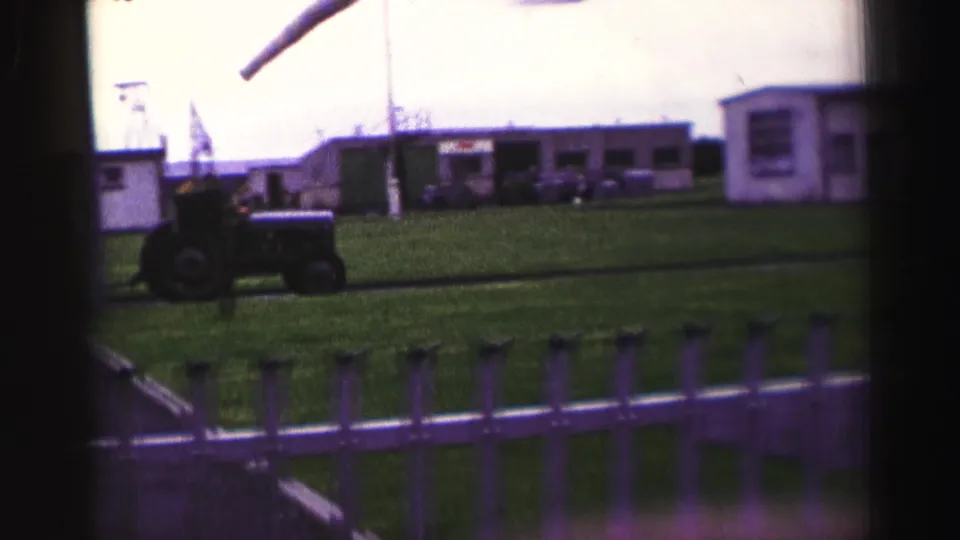 1952 United Kingdom home-movie frame showing a tractor near a standing figure, with railings, grass, and low compound buildings in view.