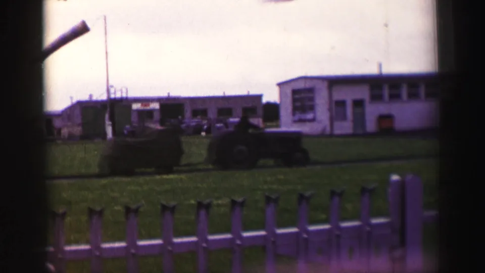 1952 United Kingdom home-movie frame showing a tractor near temporary base buildings, with white railings and grass in the foreground.