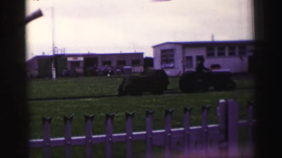 1952 United Kingdom home-movie frame showing a tractor pulling a mobile home or caravan across grass, with low buildings behind a fence.