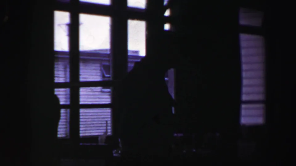 1952 United Kingdom home-movie frame looking through dark interior window frames, with a silhouetted person near the doorway or window.