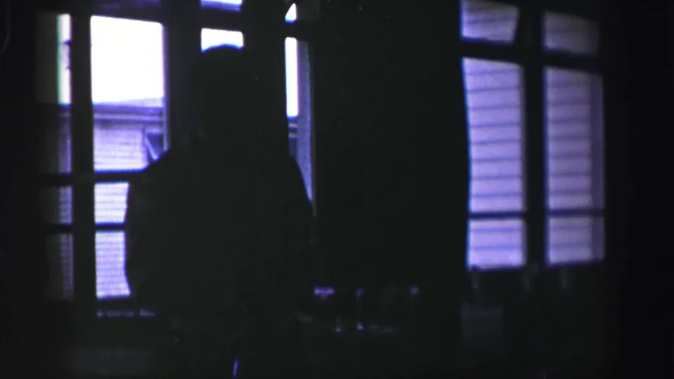 1952 United Kingdom home-movie frame with a silhouetted person near tall window panes inside a dark domestic interior.