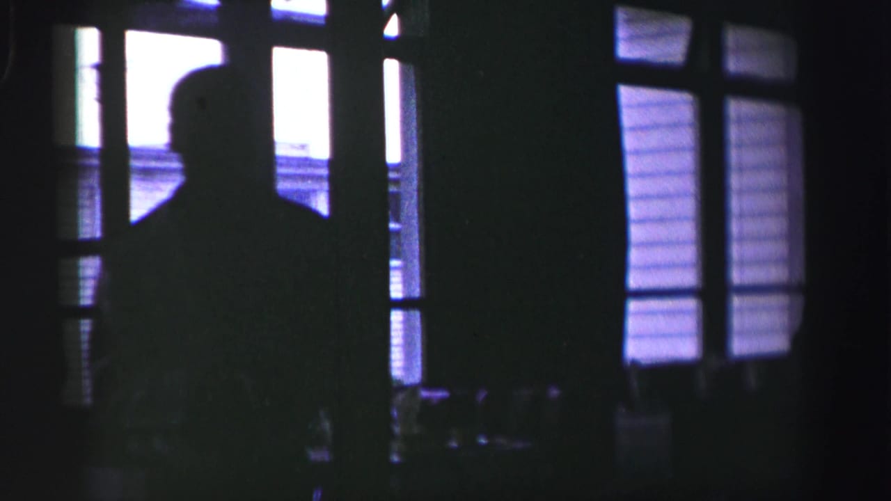Dark 8mm frame from the United Kingdom clip showing a silhouetted figure beside bright window panes at night.
