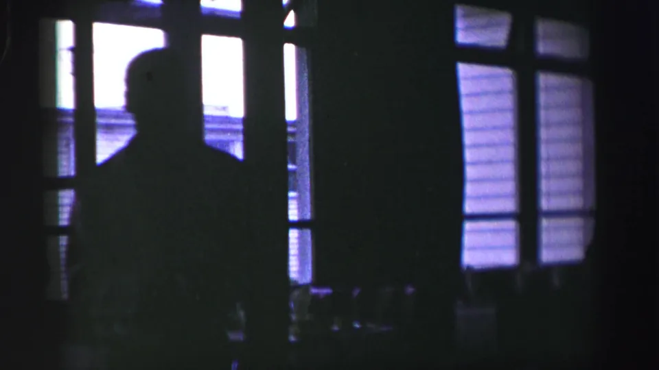 Dark 8mm frame from the United Kingdom clip showing a silhouetted figure beside bright window panes at night.
