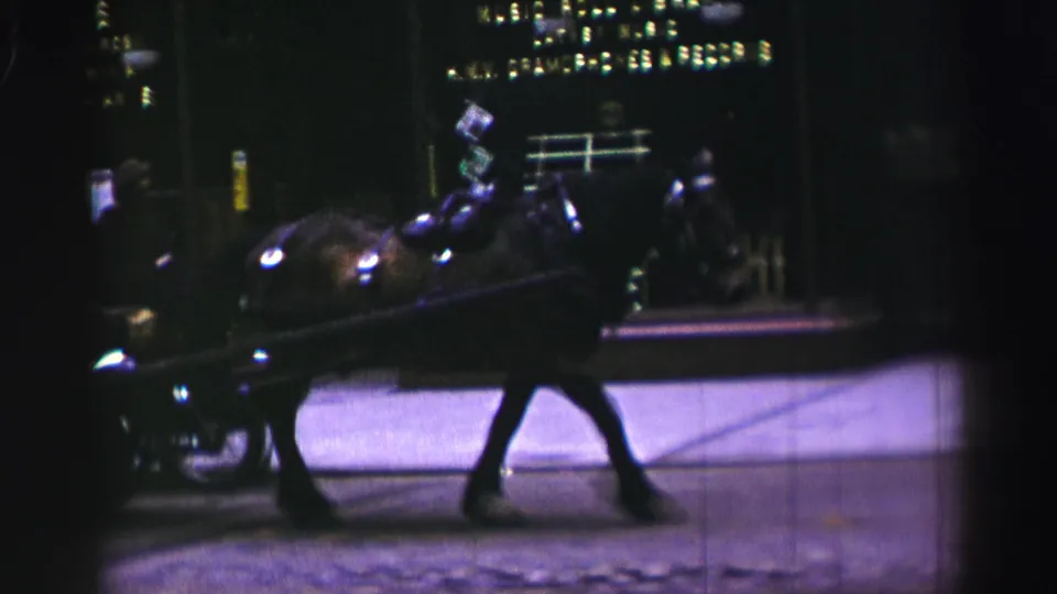 A horse-drawn cart moves along a night street in the United Kingdom, with shopfront lights and building windows behind it.