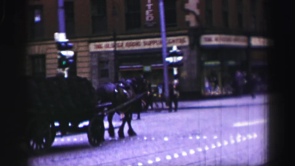 1952 United Kingdom home-movie frame showing a horse-drawn cart on a dark town street with pedestrians and lit shopfronts.