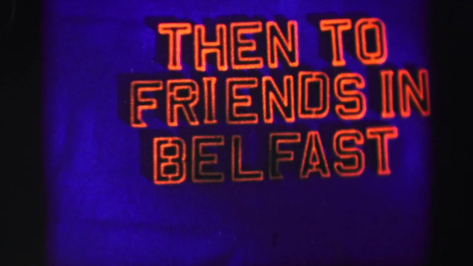 8mm frame with a glowing sign reading THEN TO FRIENDS IN BELFAST, rendered in red-orange letters on a blue-purple field.