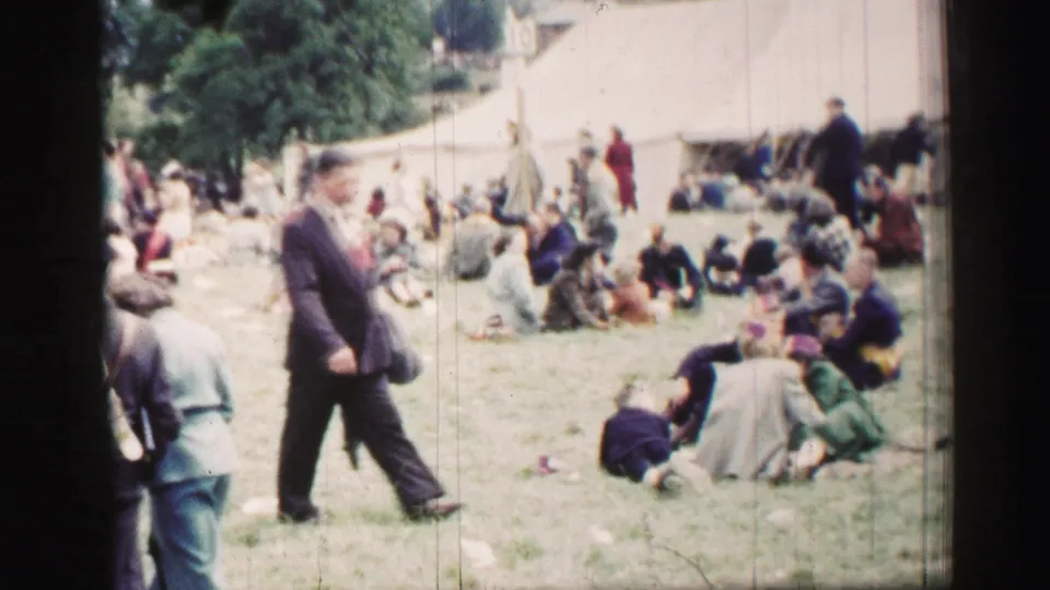 Outdoor Belfast gathering footage shows groups seated on a grassy field near a large white tent, with adults in mid-century clothing.