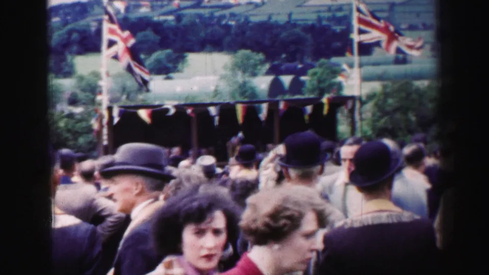 1952 United Kingdom home-movie frame showing a crowd before a bunting-covered stall with Union Jack flags and hills behind.