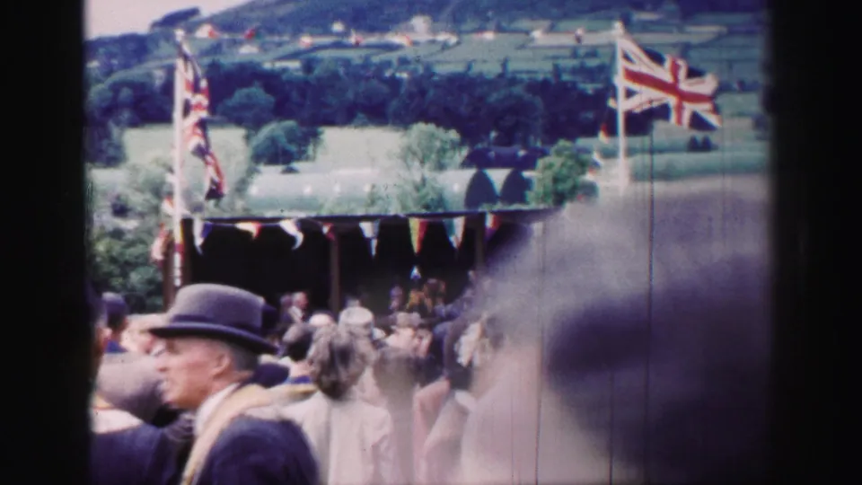 Rural United Kingdom fete crowd with Union Jack flags, partly blocked by a passerby close to the 8mm camera.