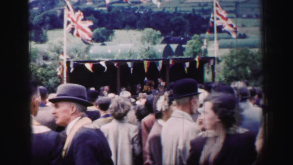 1952 United Kingdom home-movie frame with a crowd, bowler hats, bunting, Union Jack flags, and rural hills beyond the stall.
