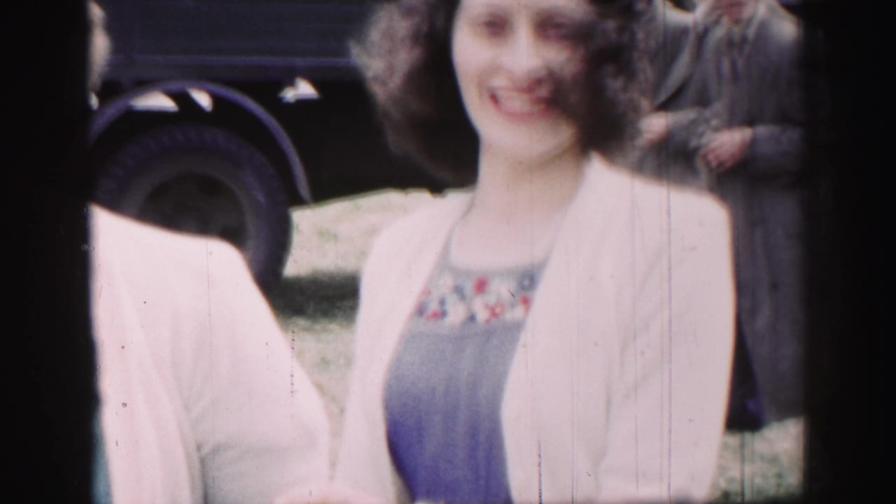 Two women stand outdoors, one in a blue dress and white sweater, with a dark vehicle edge behind them.
