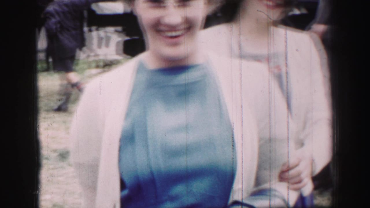 1952 United Kingdom frame of two smiling women at an outdoor gathering, one wearing a blue dress and light sweater, with figures in the background.