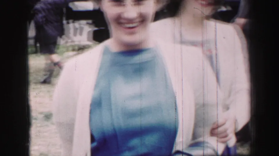 1952 United Kingdom frame of two smiling women at an outdoor gathering, one wearing a blue dress and light sweater, with figures in the background.