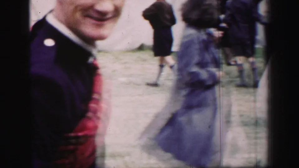 1952 United Kingdom home-movie frame with a man in dark jacket near the camera and blurred outdoor movement behind him.
