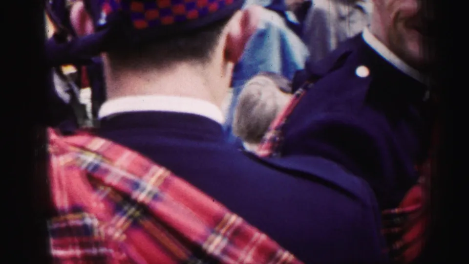 1952 United Kingdom frame showing the back of a man wearing tartan dress among people in dark jackets at an outdoor gathering.