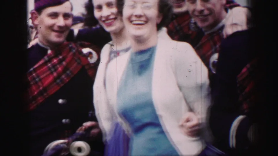 1952 United Kingdom home-movie frame of a smiling woman standing arm-in-arm near Scottish-uniformed bagpipers.