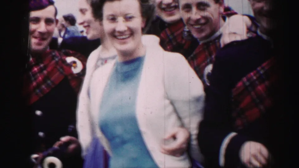 1952 United Kingdom frame of adults smiling close to the camera, including a woman beside Scottish-uniformed bagpipers.