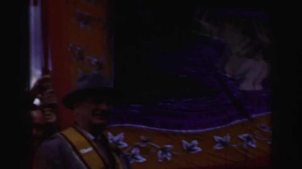 Dark handheld frame from a United Kingdom cultural gathering, with adults partly obscured beside a decorated banner or standard.