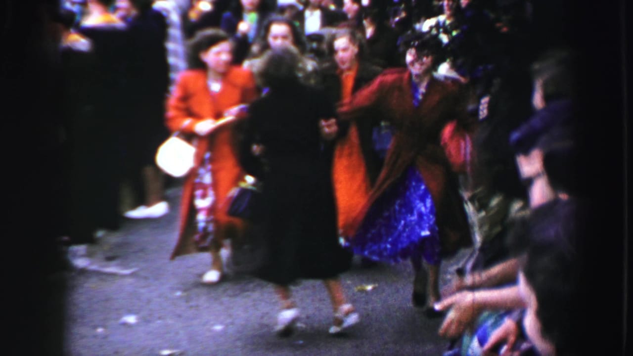 Women in colorful coats dance in a street while a crowd watches nearby.