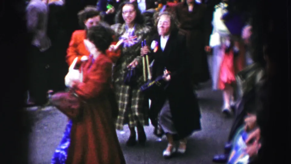 1952 United Kingdom street scene with a woman in a red coat dancing near women in plaid and dark coats among onlookers.