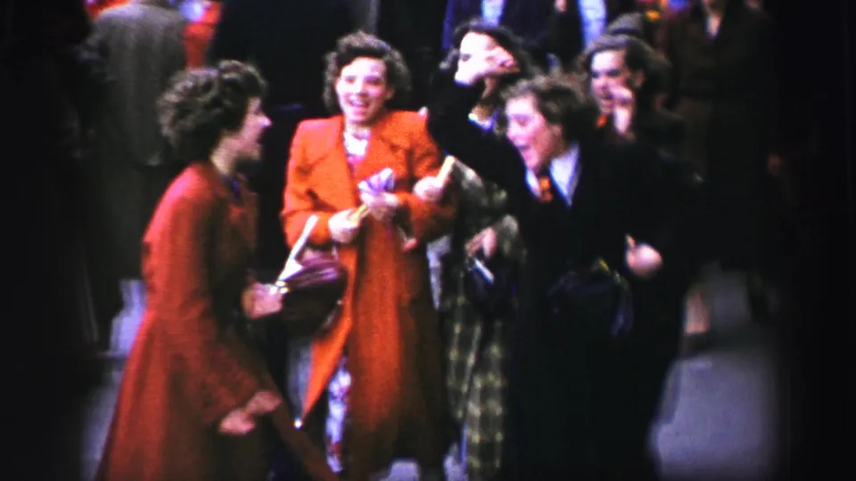 1952 United Kingdom home-movie frame of women in mid-century coats smiling and dancing together in a street crowd.