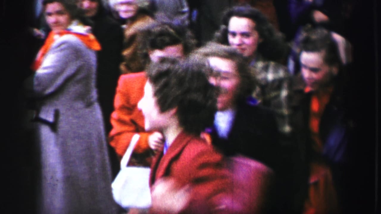 1952 United Kingdom home-movie frame showing women in colorful coats laughing and moving through a crowded street parade.