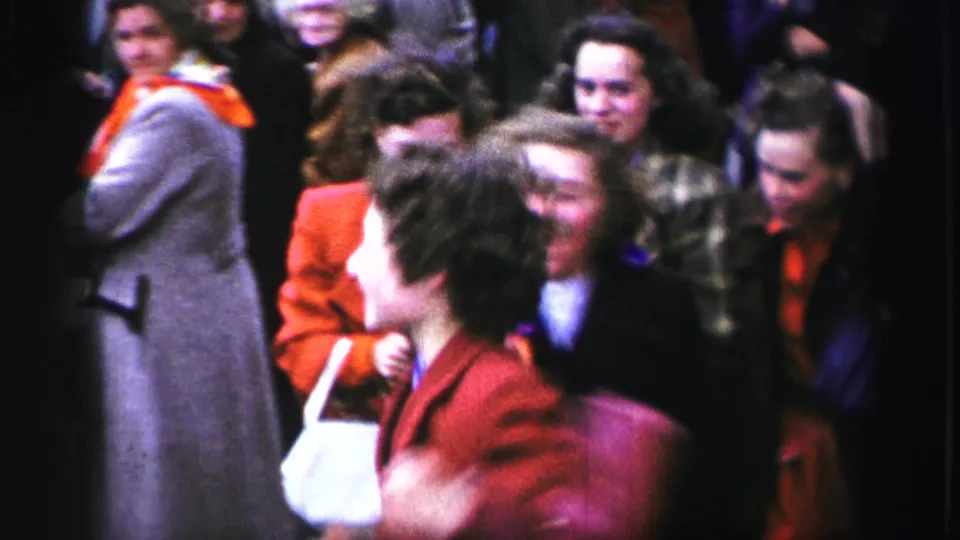 1952 United Kingdom home-movie frame showing women in colorful coats laughing and moving through a crowded street parade.
