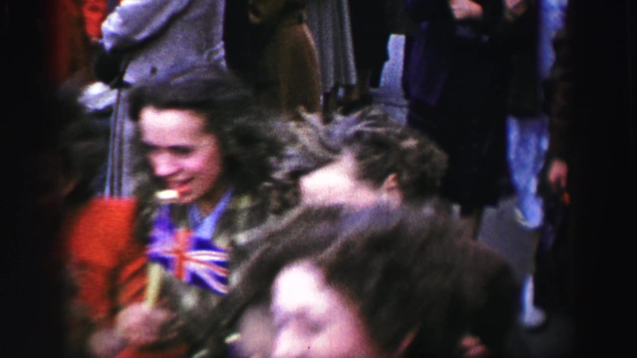 1952 United Kingdom parade frame with a smiling woman in a patterned coat near other moving spectators and participants.