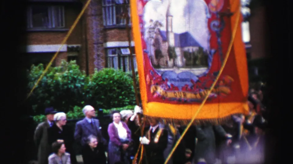 1952 United Kingdom home-movie frame showing an orange-fringed parade banner with an illustrated building scene above spectators.