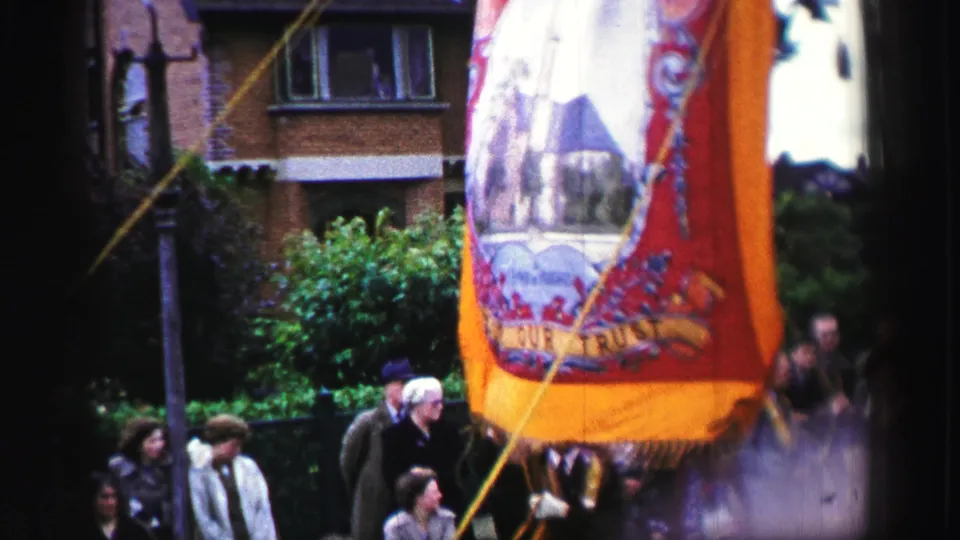 1952 United Kingdom parade frame with a decorated orange banner, support cords, spectators, brick housing, and street greenery.