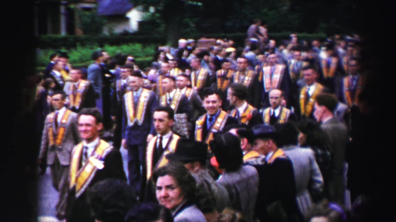 Crowd of suited men in yellow and purple sashes walking past spectators on a leafy street.