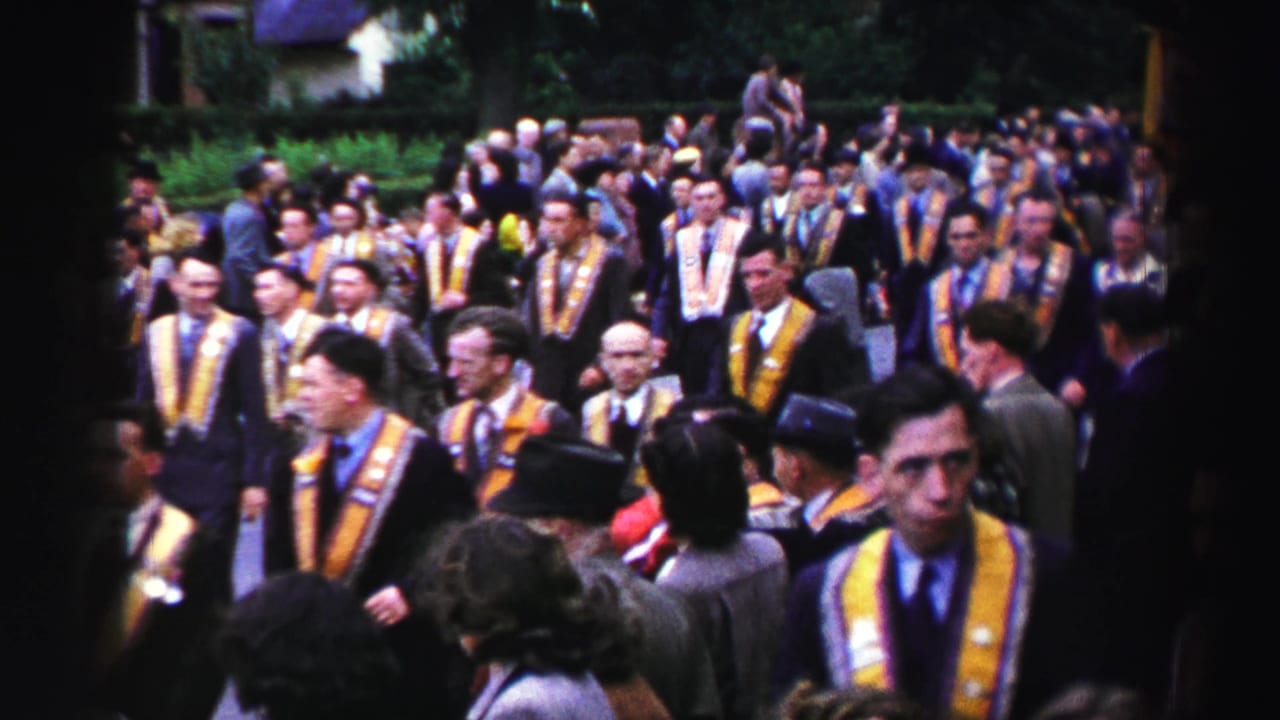 Home-movie view of a United Kingdom ceremonial procession with adult men in suits and yellow-purple sashes among spectators.
