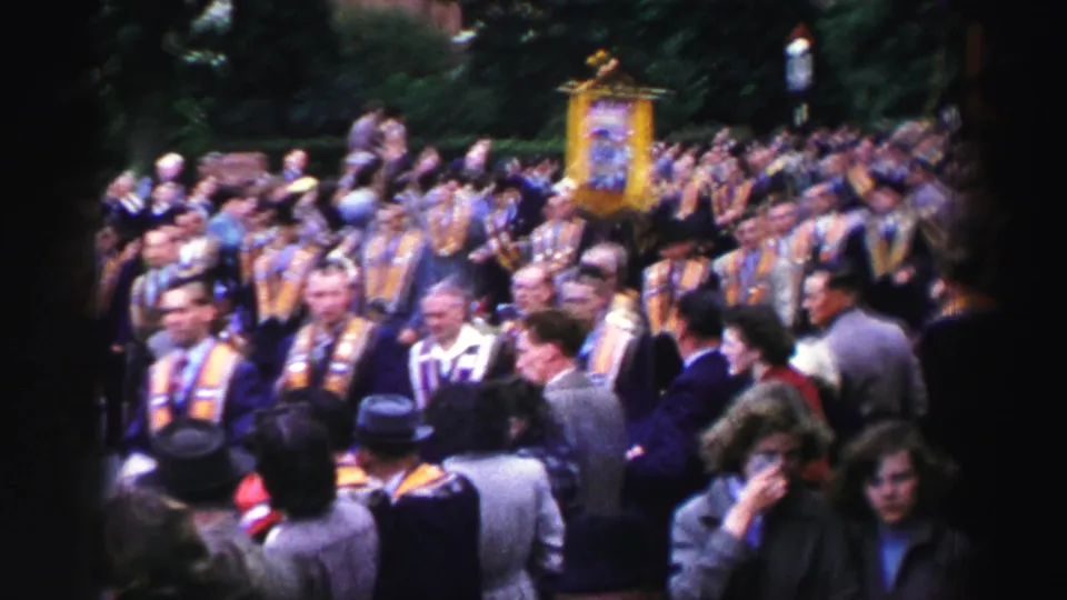 United Kingdom procession frame showing a dense crowd, many sashed men, and a yellow banner rising above the marchers.