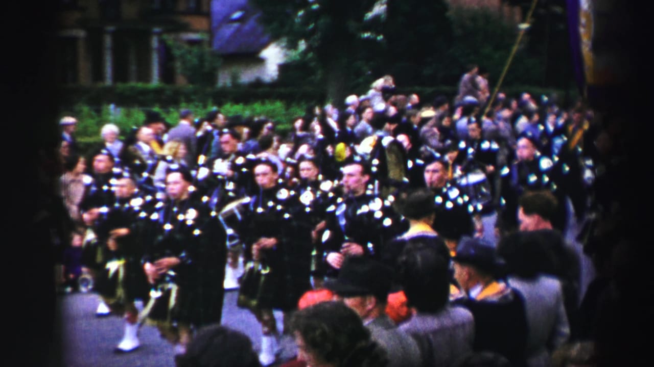 1952 United Kingdom home-movie frame showing Scottish bagpipers in kilts marching past spectators on a tree-lined street.
