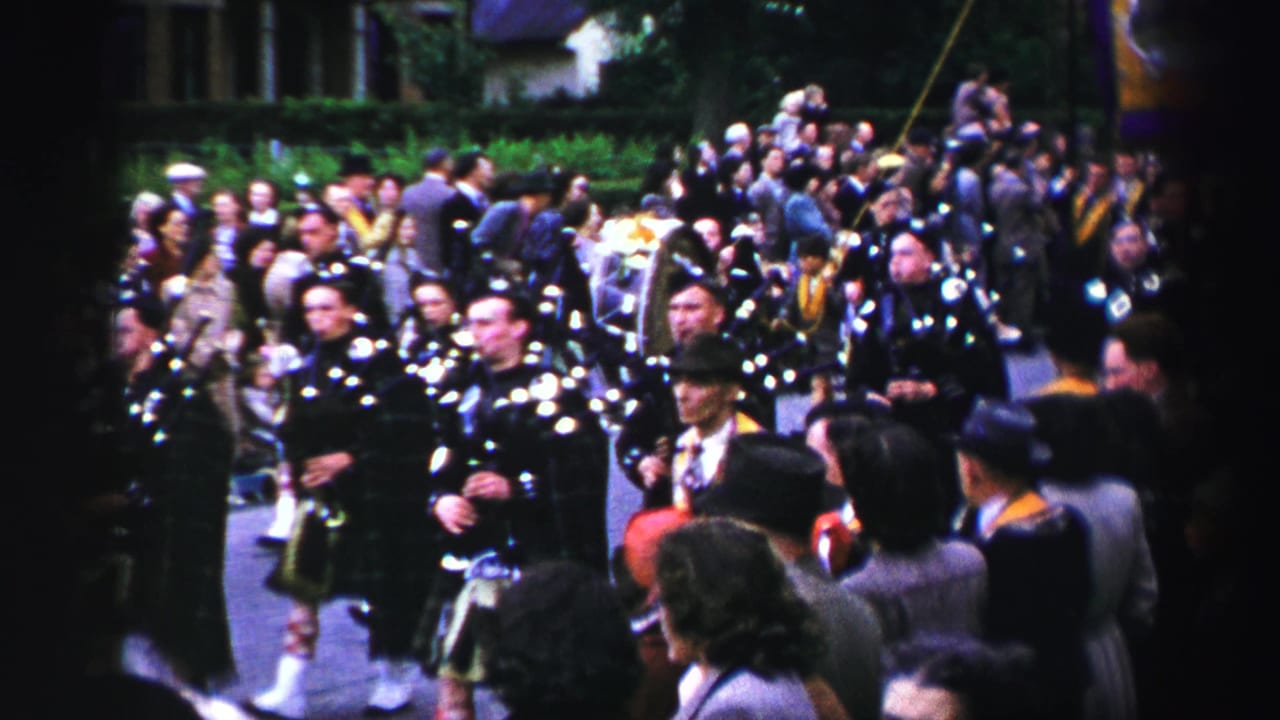 Kilted Scottish pipe band advances along a United Kingdom street, with a dense crowd and banner colors visible behind them.