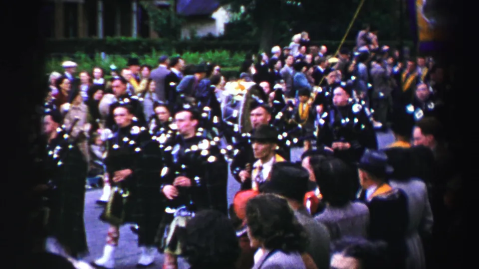 Kilted Scottish pipe band advances along a United Kingdom street, with a dense crowd and banner colors visible behind them.