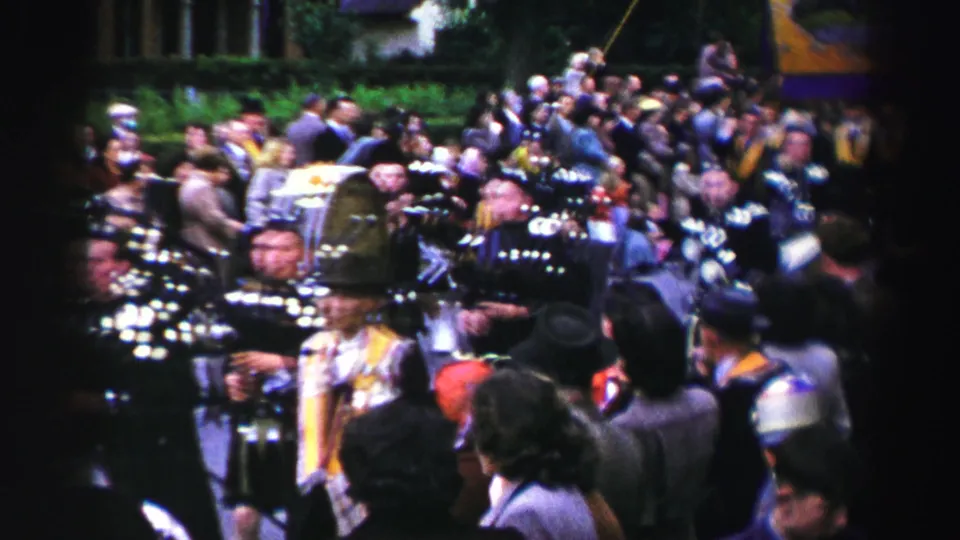 Home-movie view of the parade with kilted musicians, a nearby participant in light clothing, and spectators clustered along the street.
