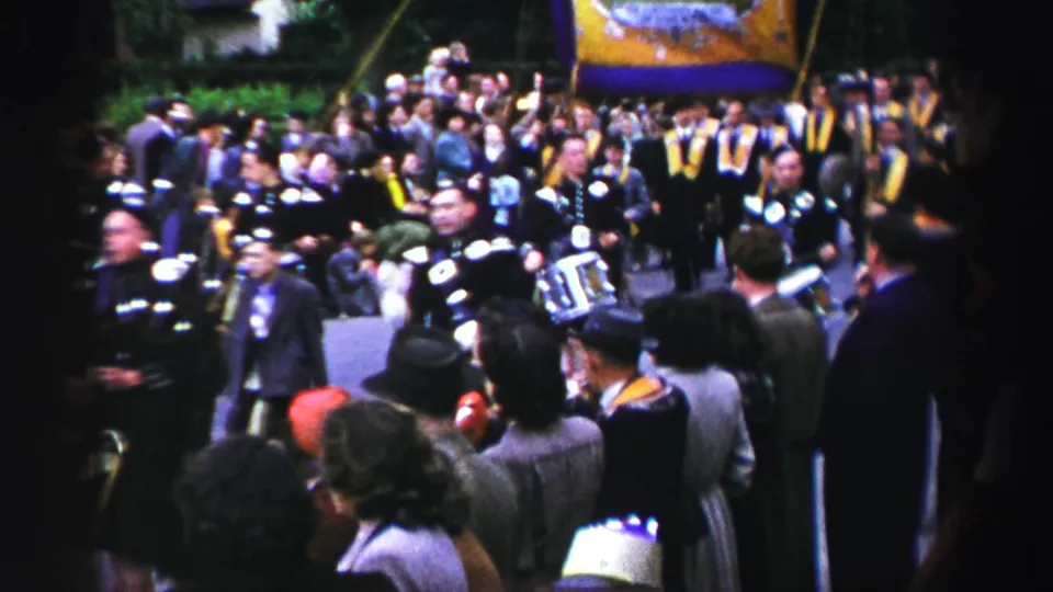 United Kingdom street parade frame with kilted musicians, clustered spectators, and a yellow and purple banner near the marching group.