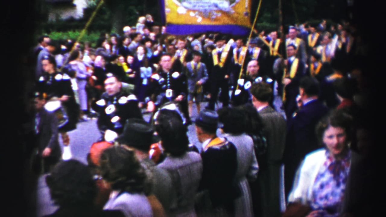 1952 United Kingdom home-movie frame showing spectators around a parade, with uniformed musicians and a large yellow banner visible.