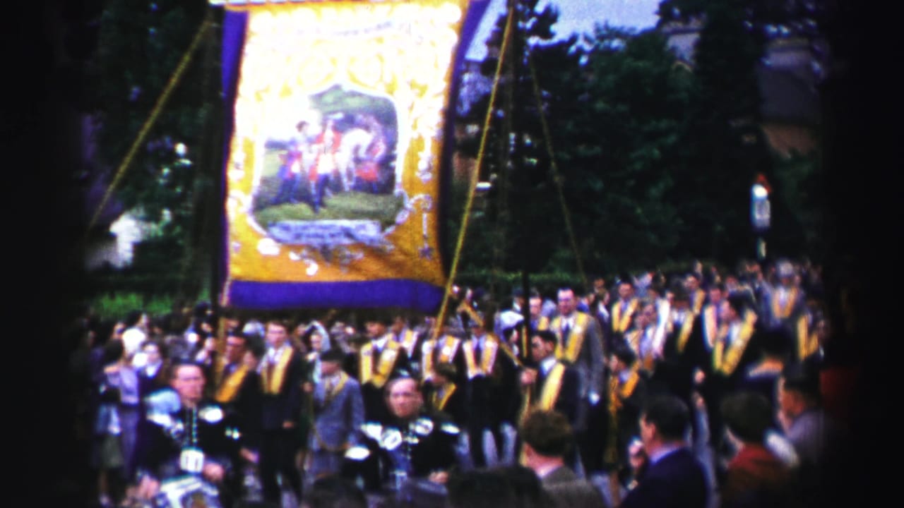 A large embroidered banner is carried through a crowded United Kingdom parade scene, with marchers in yellow sashes below.