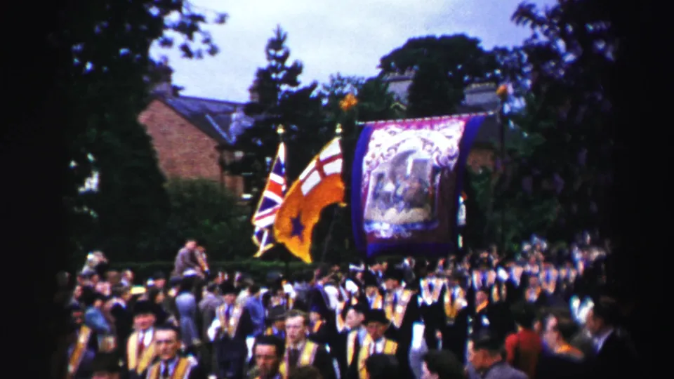 Marchers with yellow sashes, flags and a purple banner pass a crowd on a tree-lined United Kingdom street in 1952 footage.