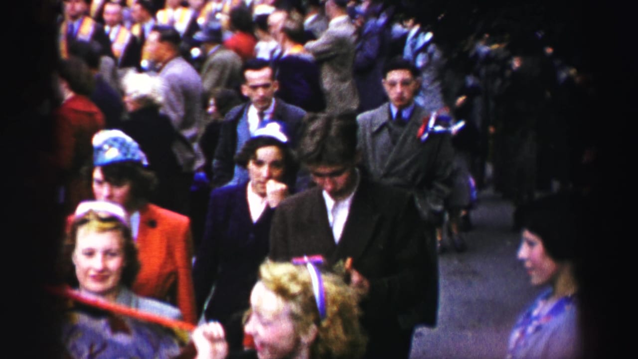 1952 United Kingdom home-movie frame showing men and women in period coats and dresses walking through a crowded parade route.