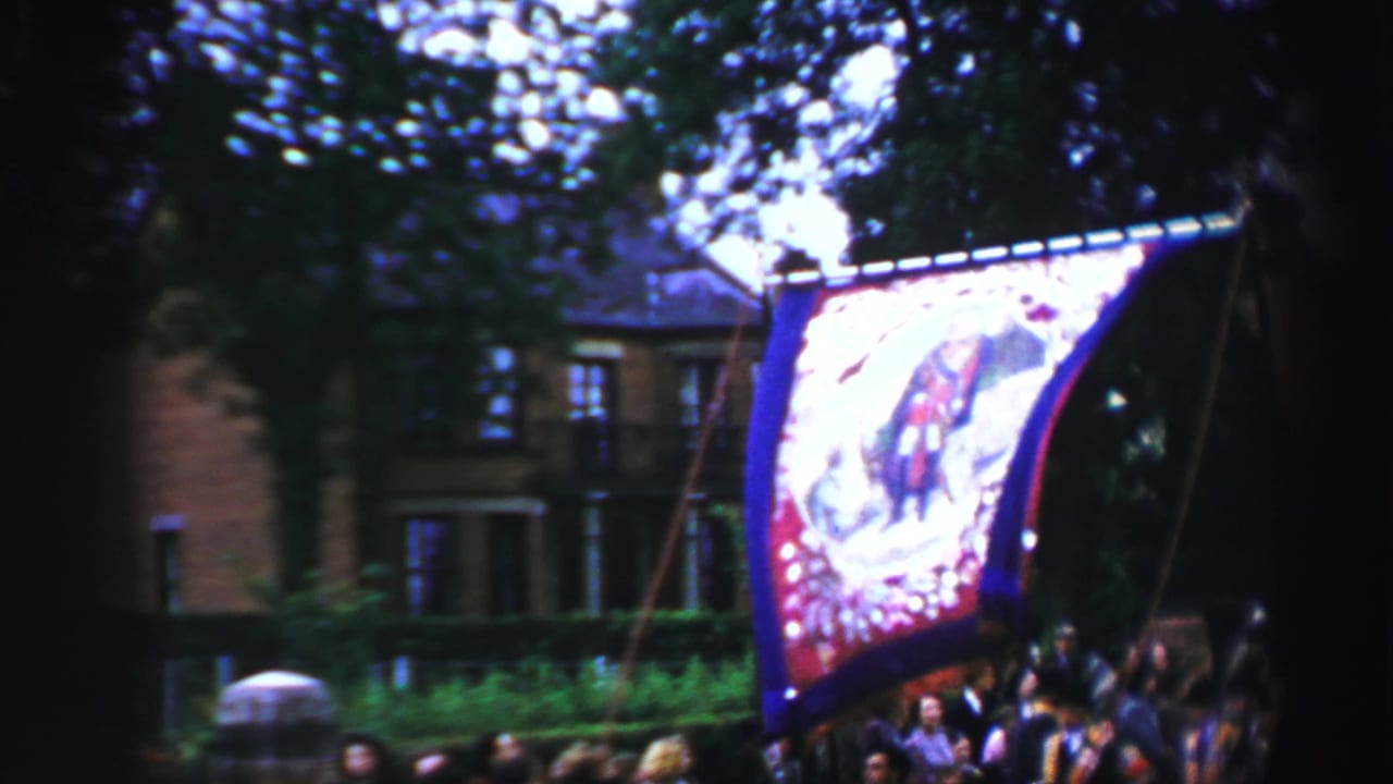 A decorated banner passes through a tree-lined United Kingdom parade route, with a building partly visible behind it.